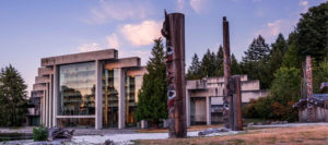 Museum of Anthropology at dusk with totem poles in the foreground and evergreens in the background. 