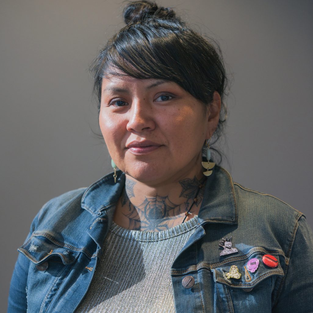 Native American with tattoos wears denim jacket with pins. Artist Melissa Cody poses for headshot style photo.