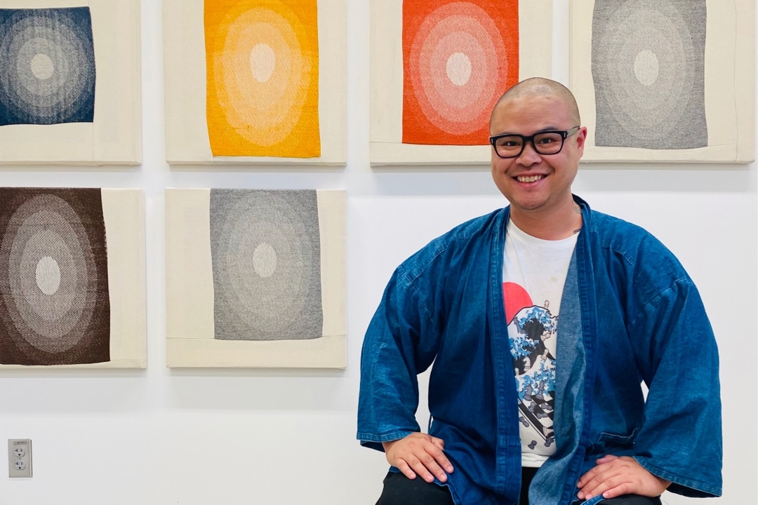 Jovencio is smiling, sitting in front of a wall display of their work.