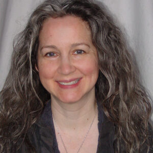 Photo portrait with long ggrey hair wearing dark grey blouse in front of light gray fabric.