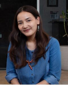 Woman with long dark hair in a blue blouse, wearing beaded earrings and necklace, smiling softly, sitting indoors in a cozy setting.